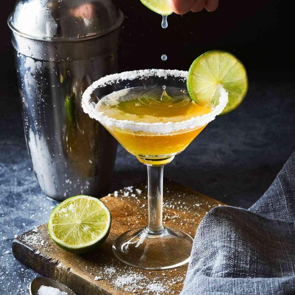 a glass of Mexican Martini served with fresh lime wedge along with cocktail mixer glass