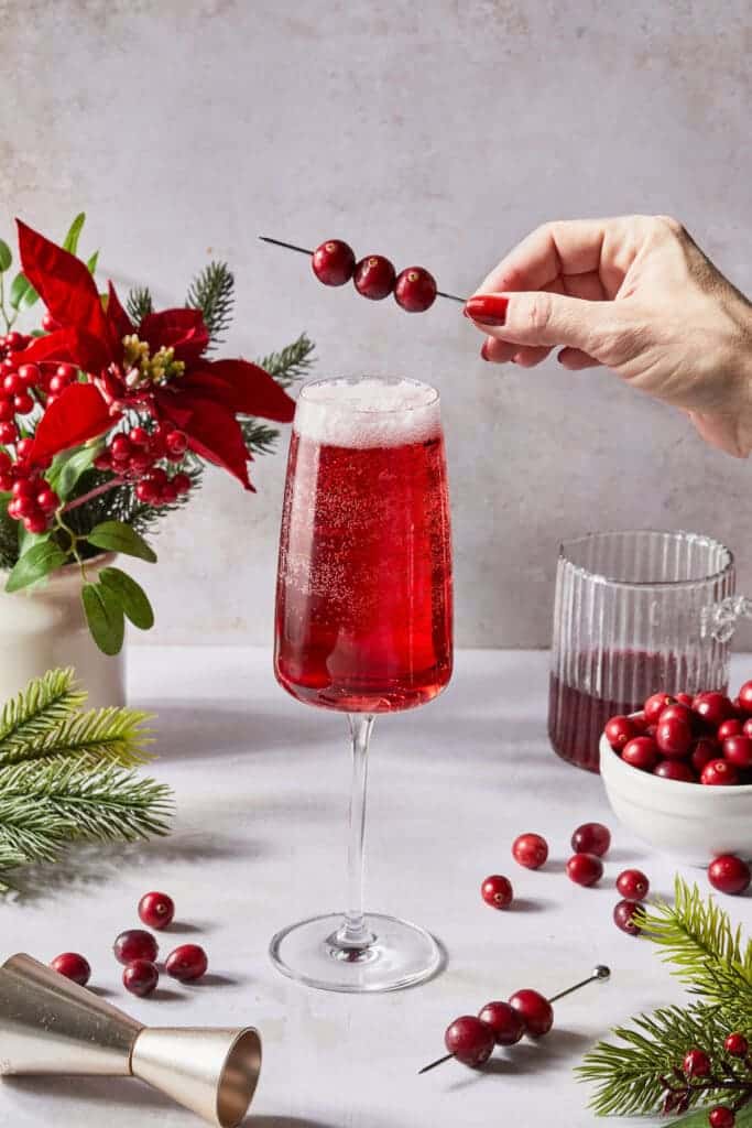 a glass of poinsettia cocktail along with cranberries on the counter and rosemerry leaves