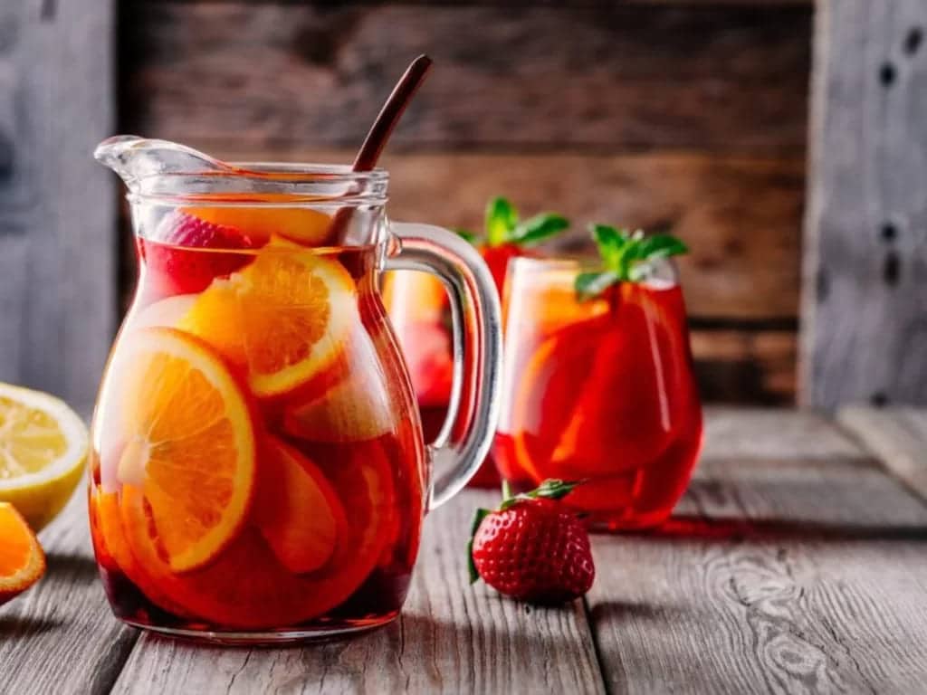 a jug of madras cocktail along with filled glasses and a strawberry orange and lemon on a counter