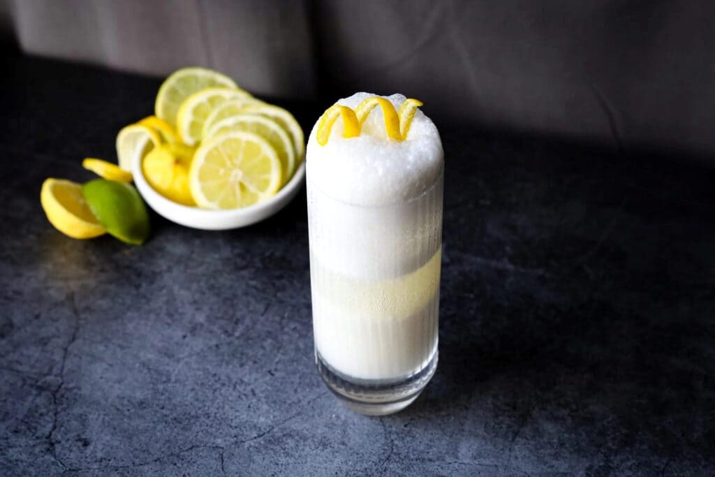 frizz drink along with the bowl full of lemon slices and frothy drink is garnish with some dried lemon peel