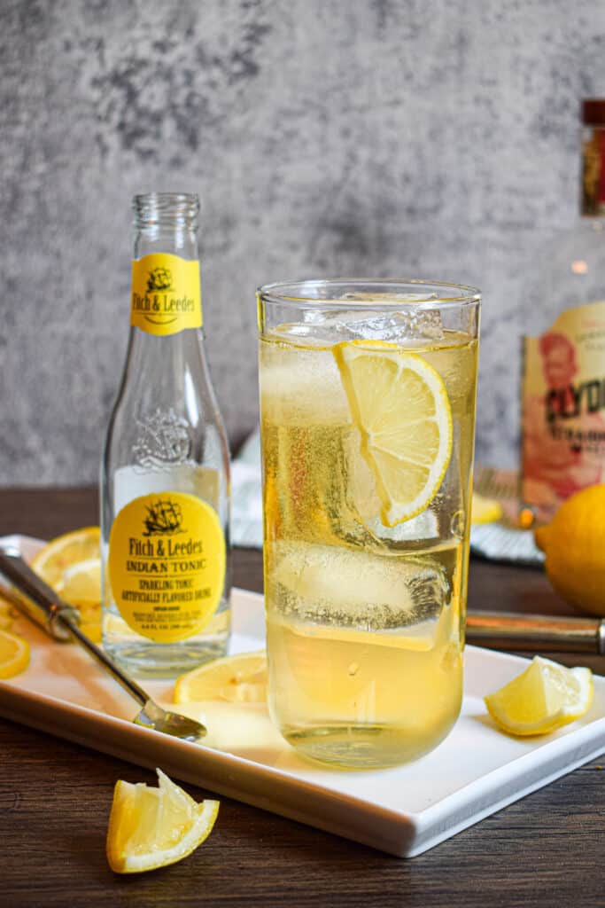 a bottle of tonic water and a glass of whiskey tonic with lemon wedge and ice