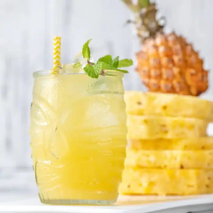 a glass of Pineapple Rum Punch garnished with mint leaves with sliced and whole pineapple in background