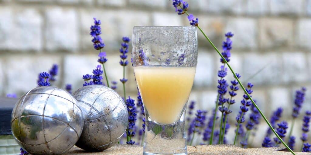 a glass of pastis placed in front of lavendar floral background and decorative balls lying nearby