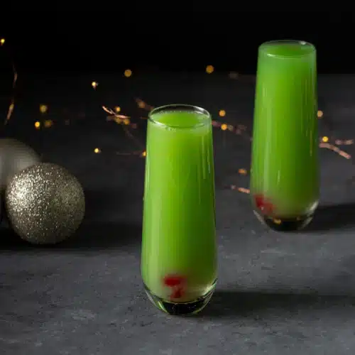 two glass of Sparkling Grinch Mimosa placed on a flat surface with balls in the background