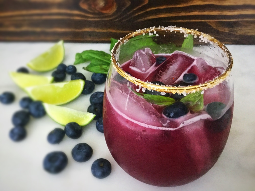 A view of a glass of a blueberry mojito with lemons and blueberries on the side